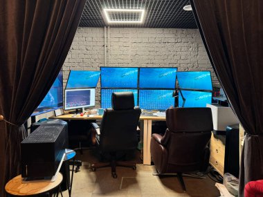 Karanlık ofisin içinde tüccar masaları var. Birden fazla monitörü olan organize bir çalışma alanı gösteriliyor. 