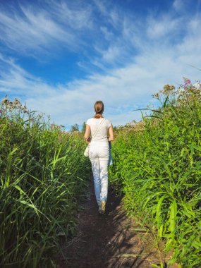 Gündüz vakti vahşi araziye giden yeşil patikada yürüyen bir kadın, turizm konsepti. Uzun otların arasında bir patika. Sabah uzun otların arasından geçen güneşli bir yol. Dikey görünüm.