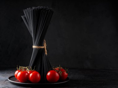 Koyu arkaplanda çiğ siyah spagetti ve kiraz domatesleri. Siyah tabakta bir demet siyah makarna. Boşluğu kopyala