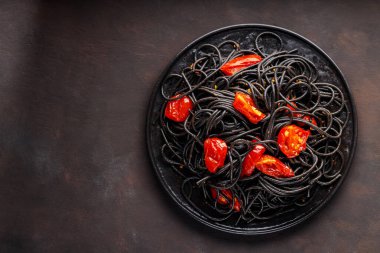 Siyah tabakta vişneli domatesli siyah spagetti. Kaynamış siyah makarna ve koyu arkaplanda kırmızı domatesler. Üst Manzara. Boşluğu kopyala