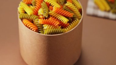 Karton bir kutuda üç renkli fusilli makarna. Bej arka planda çiğ fusilli makarna. Kamera hareketini yukarı kaldır