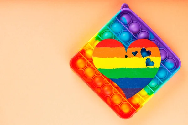 Rainbow heart and multicolored pop it fidget toys on coral background. LGBT Pride month concept. Top view. Copy space