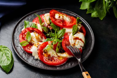 Caprese Salatası. Domates ve fesleğen yapraklı mozzarella dilimleri ve siyah tabakta çatal. Geleneksel İtalyan yemekleri