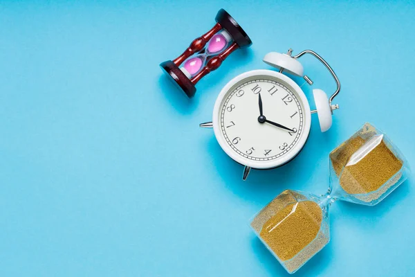 Two Hourglasses and White Alarm Clock on Blue Background, Time Concept