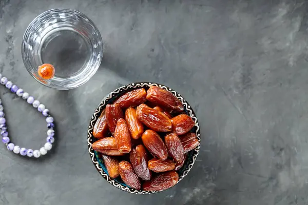 Dried Dates in Bowl, Glass of Water and Prayer Beads on Concrete, Ramadan Suhoor Concept, Copy Space, Top View