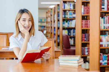 Asyalı öğrenci portresi kampüs kütüphanesinde mutlu bir gülümseme ile kendini öğrenme eğitimi üzerine bir kitap okuyor..