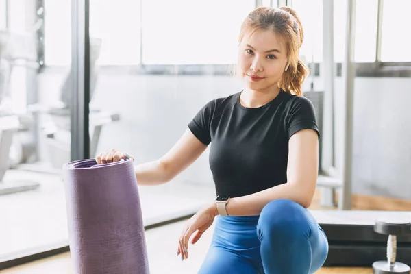 Fitness woman in sport club studio with yoga mat. Portrait happy young woman hand holding pilates mat roll at a fitness training class.