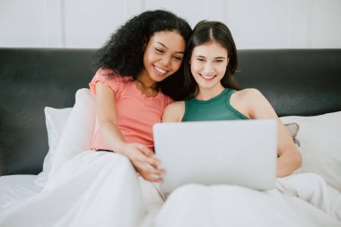 LGBT kadınları yatakta oturup dizüstü bilgisayar izliyorlar. Mutlu bir şekilde gülümsüyorlar. Ev tatili aktivitelerinde birlikte yaşıyorlar.