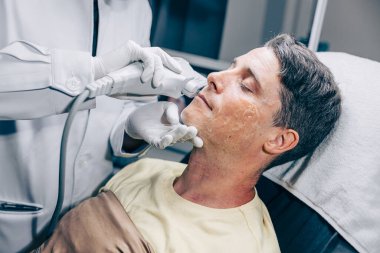 Yaşlanma karşıtı klinikte kırışıklıkları ve yaraları iyileştirmek için yetişkin bir erkekle yüz bakımı teknolojisi kullanan cilt doktoru.