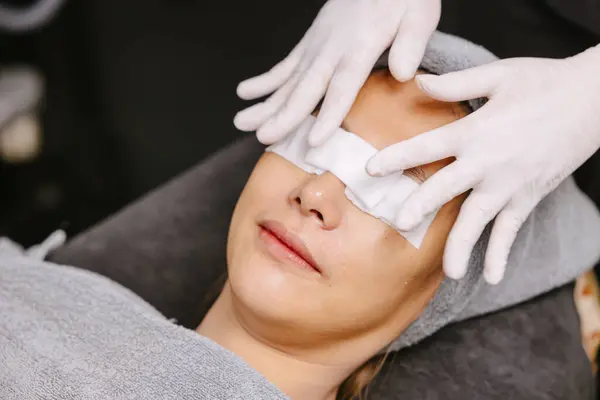 Anti-aging clinic woman closed eyes with cotton pad enjoying skincare procedure. Masseuse gentle massage client face and eyes