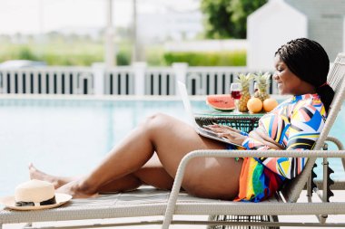 Afrikalı siyahi bir kadın yaz tatilinde bilgisayar kumandası kullanıyor. Lüks otel havuzunda dinleniyor. Serbest bikinili kadın internet havuz başında çalışıyor..