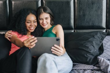 LGBT kadınları yatakta oturup tablet ekranı izlerken mutlu bir şekilde gülümsüyor ve evde tatil aktivitelerinde birlikte yaşıyorlar.