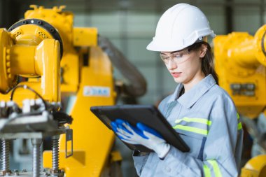Makine fabrikasında çalışan mühendis kadın. Kadın mühendis, üretim tesisinde robot kolu tabletle kontrol ediyor.