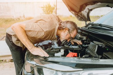 Yol kenarında arabası kırılmış yaşlı bir erkek sorun noktası kontrolü ve tanımlaması için motor altına bakıyor.