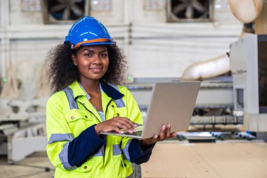 Güvenlik elbisesi olan Afrikalı siyahi kadın işçi, ahşap atölye mobilya fabrikasında dizüstü bilgisayar programı işletim makinesi kullanıyor.