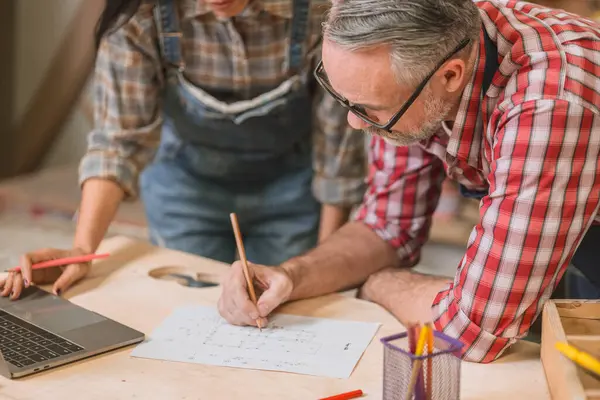 Profesyonel kıdemli marangoz kadın takımıyla çalışıyor fabrikadaki ahşap atölyede ahşap mobilya tasarımı yapıyor.