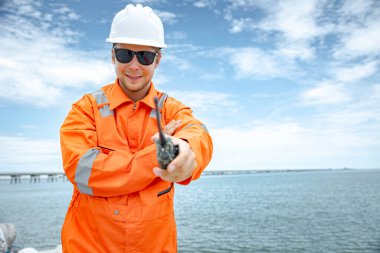 Profesyonel portre mutlu gülümseyen petrol işçisi kasketli erkek, okyanusa ulaşan doğal gaz borusuna karşı duruyor.