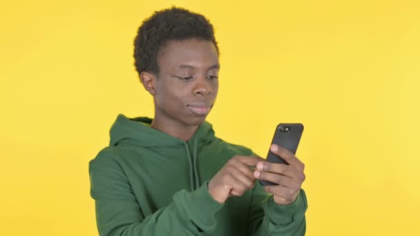 黄色の背景にスマートフォンで祝うカジュアルアフリカ人男性 — ストック動画