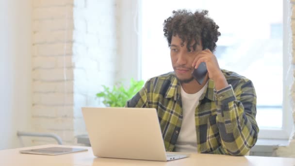 ラップトップを使用している間に電話で話している若いアフリカ人男性 — ストック動画