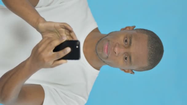 青の背景にスマートフォンを閲覧する若いアフリカ人男性の垂直ビデオ — ストック動画