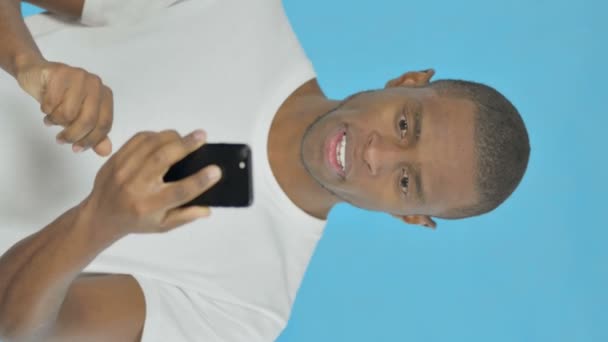 青を背景にスマートフォンで祝う若いアフリカ人男性の垂直ビデオ — ストック動画