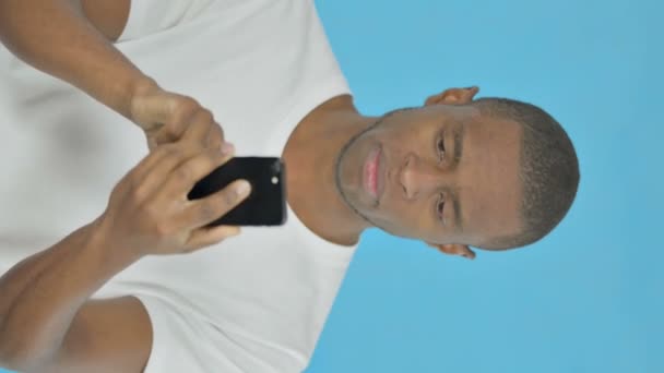 青の背景にスマートフォン上の若いアフリカの男の損失の垂直ビデオ — ストック動画