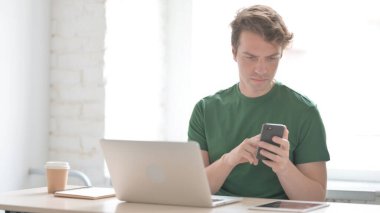 Gelişigüzel genç adam dizüstü bilgisayar kullanırken akıllı telefon kullanıyor