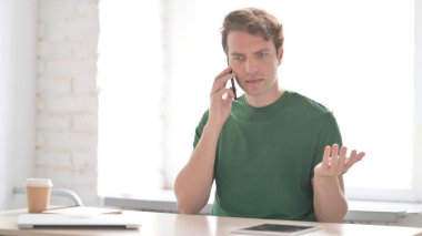 Kızgın Gündelik Genç Adam İş yerinde Akıllı Telefonla Konuşuyor