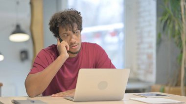 Young African Man Talking on Phone while using Laptop