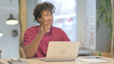 Young African Man Pointing at Camera while using Laptop