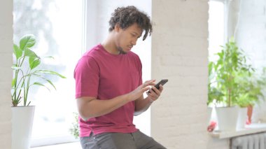 Young African Man using Smartphone while Sitting in Window