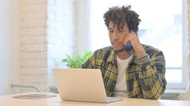 Young African Man Talking on Phone while using Laptop