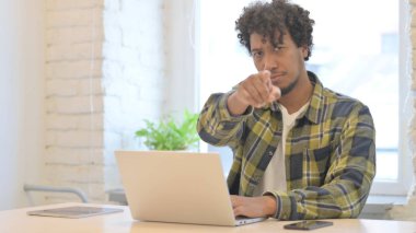 Young African Man Pointing at Camera while using Laptop