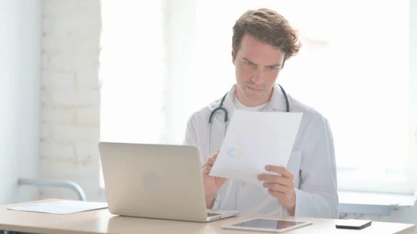 Male Doctor Working on Medical Report and Laptop in Clinic