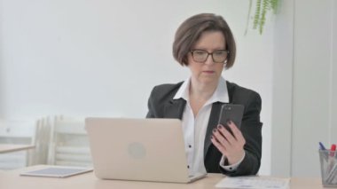 Old Senior Businesswoman using Smartphone while using Laptop
