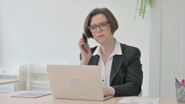 Displeased Old Senior Businesswoman Talking Angrily on Smartphone at Work