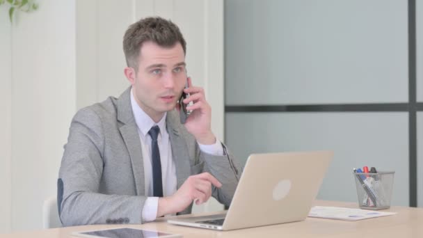 Jovem Empresário Falando Por Telefone Enquanto Usa Laptop — Vídeo de Stock