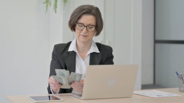 Old Senior Businesswoman Counting Dollar while Working on Laptop