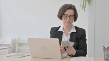 Old Senior Businesswoman Counting Dollar while Working on Laptop