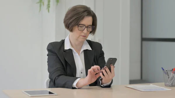 资深商界妇女在办公室的智能手机上浏览互联网 — 图库照片