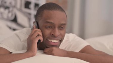 Yatakta karın üstü yatarken telefonda konuşan Afrikalı adam.