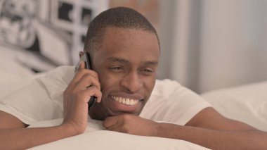 Yatakta karın üstü yatarken telefonda konuşan Afrikalı adam.