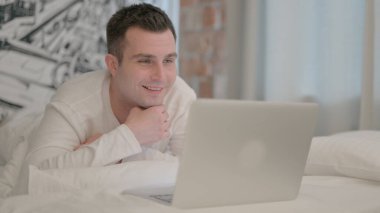 Dizüstü bilgisayarda görüntülü konuşma yapan genç adam yatakta karın üstü yatarken...