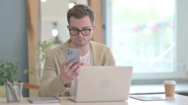 Dizüstü bilgisayarda çalışan ve akıllı telefon kullanan genç bir iş adamı.