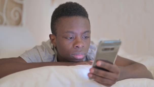 胃の上に横たわっている間 スマートフォンを使用して若いアフリカ人男性 — ストック動画