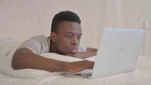 ベッドで胃の上に横たわっている間 ラップトップで働く若いアフリカ人男性 — ストック動画