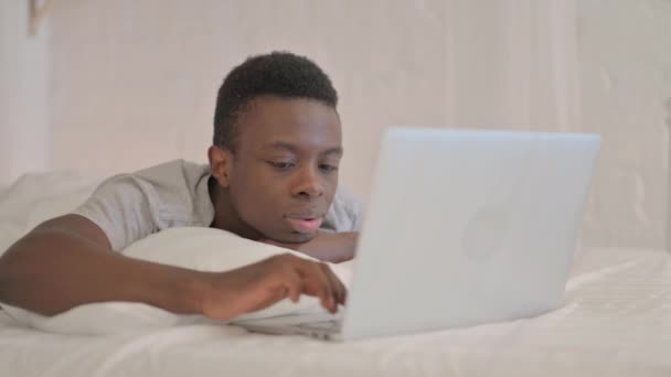 ベッドで胃の上に横たわっている間 ラップトップ上の損失によってショックを受けた若いアフリカ人男性 — ストック動画