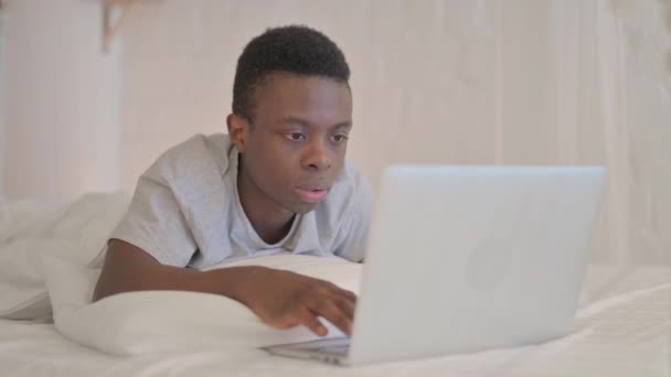 ベッドで胃の上に横たわっている間 ラップトップ上で成功を祝う若いアフリカ人男性 — ストック動画