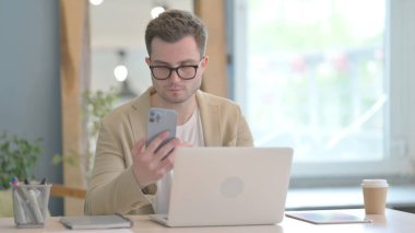 Dizüstü bilgisayarda çalışan ve akıllı telefon kullanan genç bir iş adamı.