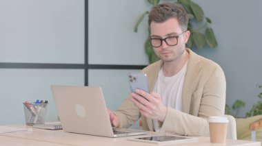 Dizüstü bilgisayarda çalışan ve akıllı telefon kullanan genç bir iş adamı.